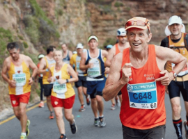 A group of marathon runners, wearing race bibs and colorful athletic gear, run on a winding road beside a rocky hillside. The man in front, smiling, wears an orange tank top and blue bib labeled C648.