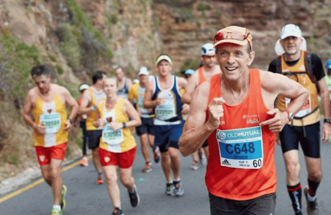 A group of marathon runners, wearing race bibs and colorful athletic gear, run on a winding road beside a rocky hillside. The man in front, smiling, wears an orange tank top and blue bib labeled C648.