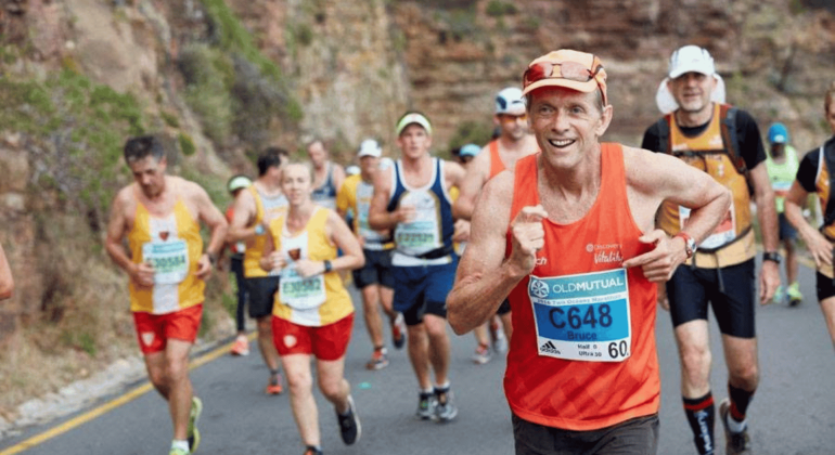 A group of marathon runners, wearing race bibs and colorful athletic gear, run on a winding road beside a rocky hillside. The man in front, smiling, wears an orange tank top and blue bib labeled C648.