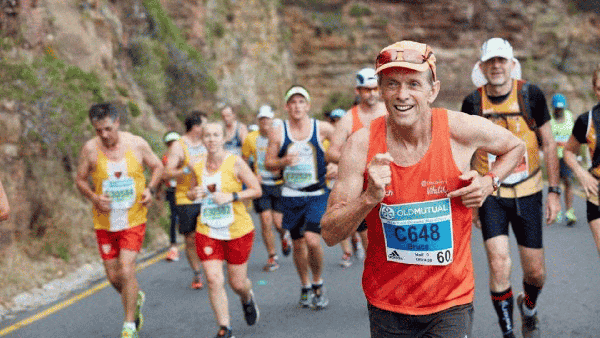 A group of marathon runners, wearing race bibs and colorful athletic gear, run on a winding road beside a rocky hillside. The man in front, smiling, wears an orange tank top and blue bib labeled C648.