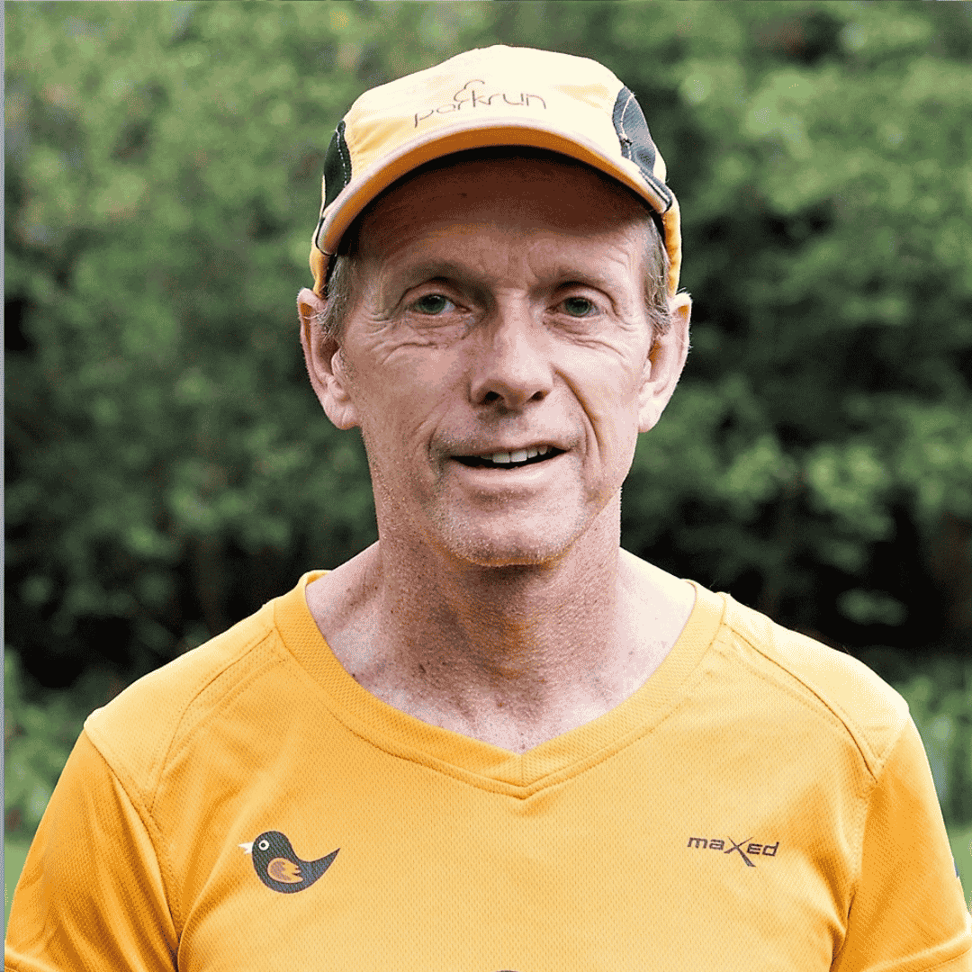 A middle-aged man wearing a yellow parkrun cap and matching yellow shirt stands outdoors in front of green foliage, smiling at the camera.