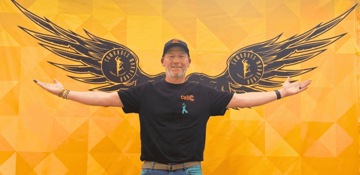 A man stands smiling with arms outstretched in front of a yellow wall painted with large black wings, giving the appearance that he has wings. He wears a black shirt, cap, and blue awareness ribbon.
