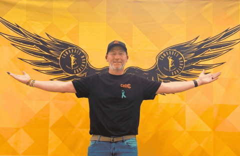 A man stands smiling with arms outstretched in front of a yellow wall painted with large black wings, giving the appearance that he has wings. He wears a black shirt, cap, and blue awareness ribbon.