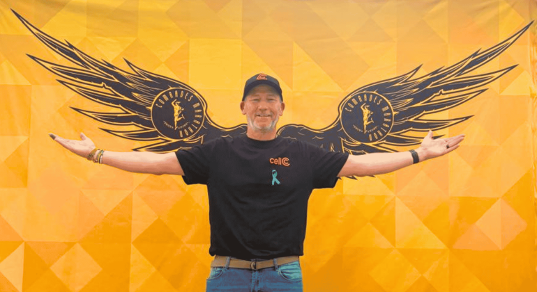 A man stands smiling with arms outstretched in front of a yellow wall painted with large black wings, giving the appearance that he has wings. He wears a black shirt, cap, and blue awareness ribbon.