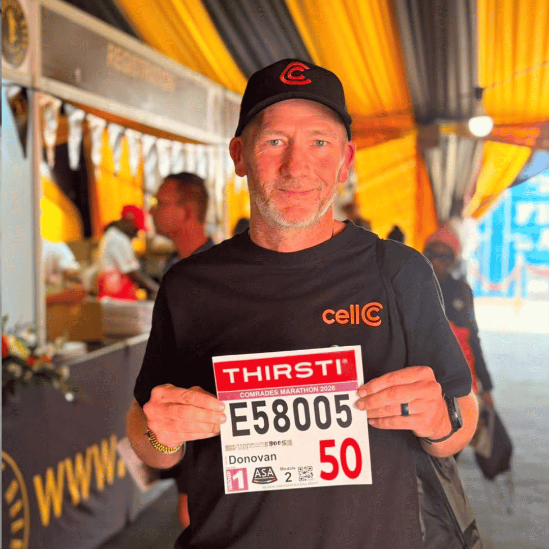 A man wearing a black Cell C shirt and cap stands indoors, holding a Comrades Marathon race number labeled “E58005” with “Donovan” and the number 50, under a yellow-and-orange tent.