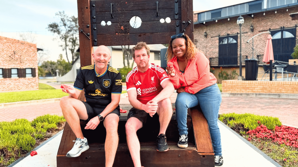 Three people smile and pose on a large wooden stock structure outdoors. Two men wear soccer jerseys, and a woman in a red top leans in from the right, channeling Ali Campbell's laid-back vibe. A brick building and greenery appear in the background.