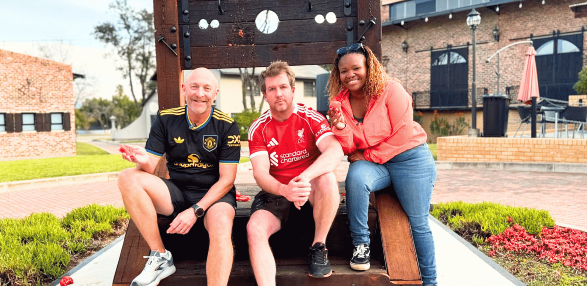 Three people smile and pose on a large wooden stock structure outdoors. Two men wear soccer jerseys, and a woman in a red top leans in from the right, channeling Ali Campbell's laid-back vibe. A brick building and greenery appear in the background.