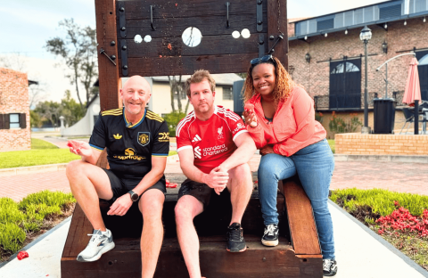 Three people smile and pose on a large wooden stock structure outdoors. Two men wear soccer jerseys, and a woman in a red top leans in from the right, channeling Ali Campbell's laid-back vibe. A brick building and greenery appear in the background.