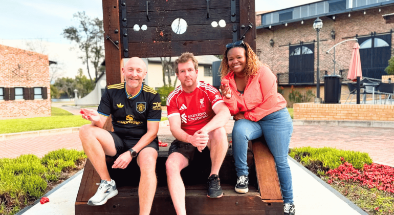 Three people smile and pose on a large wooden stock structure outdoors. Two men wear soccer jerseys, and a woman in a red top leans in from the right, channeling Ali Campbell's laid-back vibe. A brick building and greenery appear in the background.
