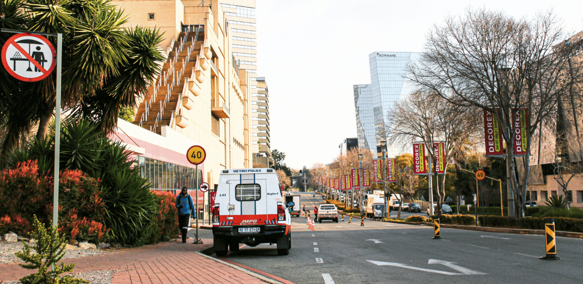 A city street with buildings, trees, and banners; a Metro Police van is parked by the sidewalk, and a &ldquo;no pedestrian crossing&rdquo; sign is visible in the foreground. Traffic cones line part of the road.