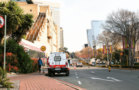 A city street with buildings, trees, and banners; a Metro Police van is parked by the sidewalk, and a &ldquo;no pedestrian crossing&rdquo; sign is visible in the foreground. Traffic cones line part of the road.