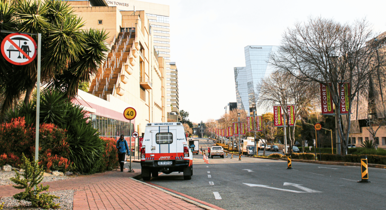 A city street with buildings, trees, and banners; a Metro Police van is parked by the sidewalk, and a “no pedestrian crossing” sign is visible in the foreground. Traffic cones line part of the road.
