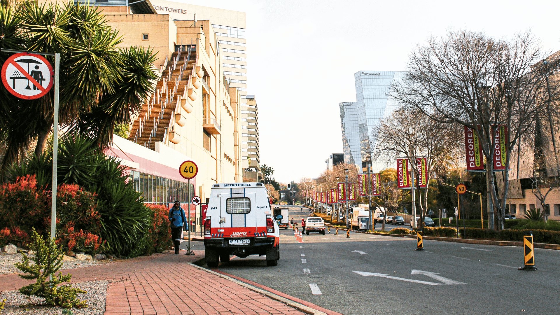 A city street with buildings, trees, and banners; a Metro Police van is parked by the sidewalk, and a “no pedestrian crossing” sign is visible in the foreground. Traffic cones line part of the road.