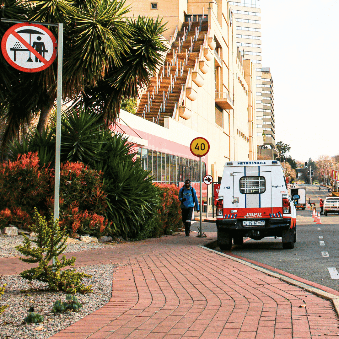 A Metro Police van is parked on a city street next to a sidewalk with plants. A person walks nearby, and a sign prohibits sitting or lying on the pavement. Tall buildings and a 40 km/h speed limit sign are visible.