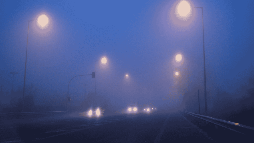 Cars drive on a foggy road at night, illuminated by streetlights. Headlights and streetlights create glowing orbs in the dense blue mist, making the surroundings appear blurry and obscured.