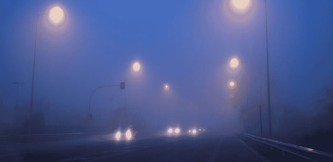 Cars drive on a foggy road at night, illuminated by streetlights. Headlights and streetlights create glowing orbs in the dense blue mist, making the surroundings appear blurry and obscured.