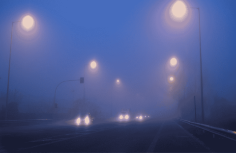 Cars drive on a foggy road at night, illuminated by streetlights. Headlights and streetlights create glowing orbs in the dense blue mist, making the surroundings appear blurry and obscured.