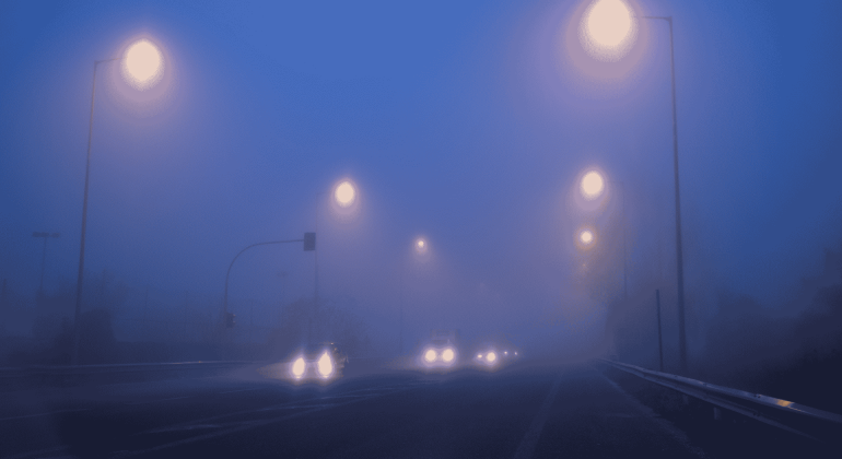 Cars drive on a foggy road at night, illuminated by streetlights. Headlights and streetlights create glowing orbs in the dense blue mist, making the surroundings appear blurry and obscured.