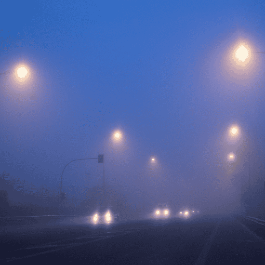 Cars drive down a street at night in heavy fog, with streetlights glowing softly and headlights illuminating the misty air. The scene appears quiet and atmospheric with low visibility.