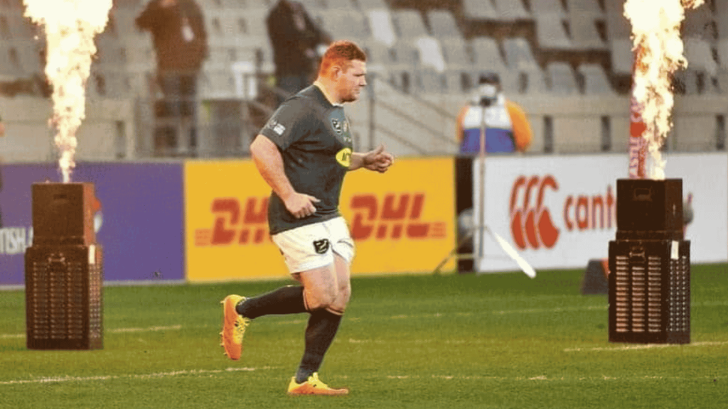 A rugby player in uniform runs onto the field between two flame machines, with empty stadium seats and advertising boards visible in the background.