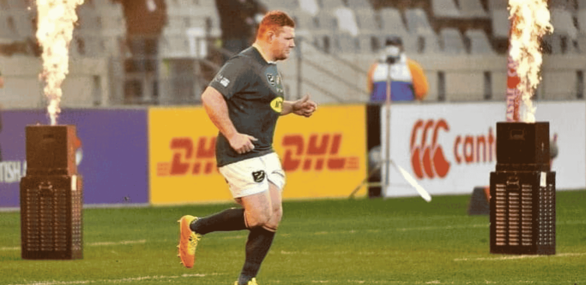 A rugby player in uniform runs onto the field between two flame machines, with empty stadium seats and advertising boards visible in the background.