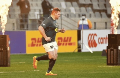 A rugby player in uniform runs onto the field between two flame machines, with empty stadium seats and advertising boards visible in the background.