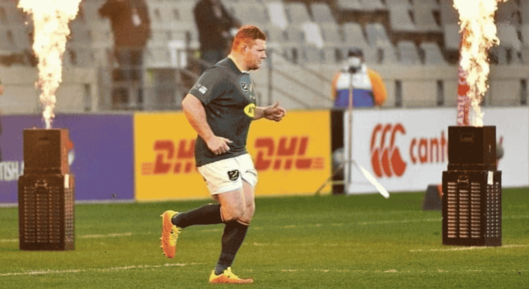 A rugby player in uniform runs onto the field between two flame machines, with empty stadium seats and advertising boards visible in the background.