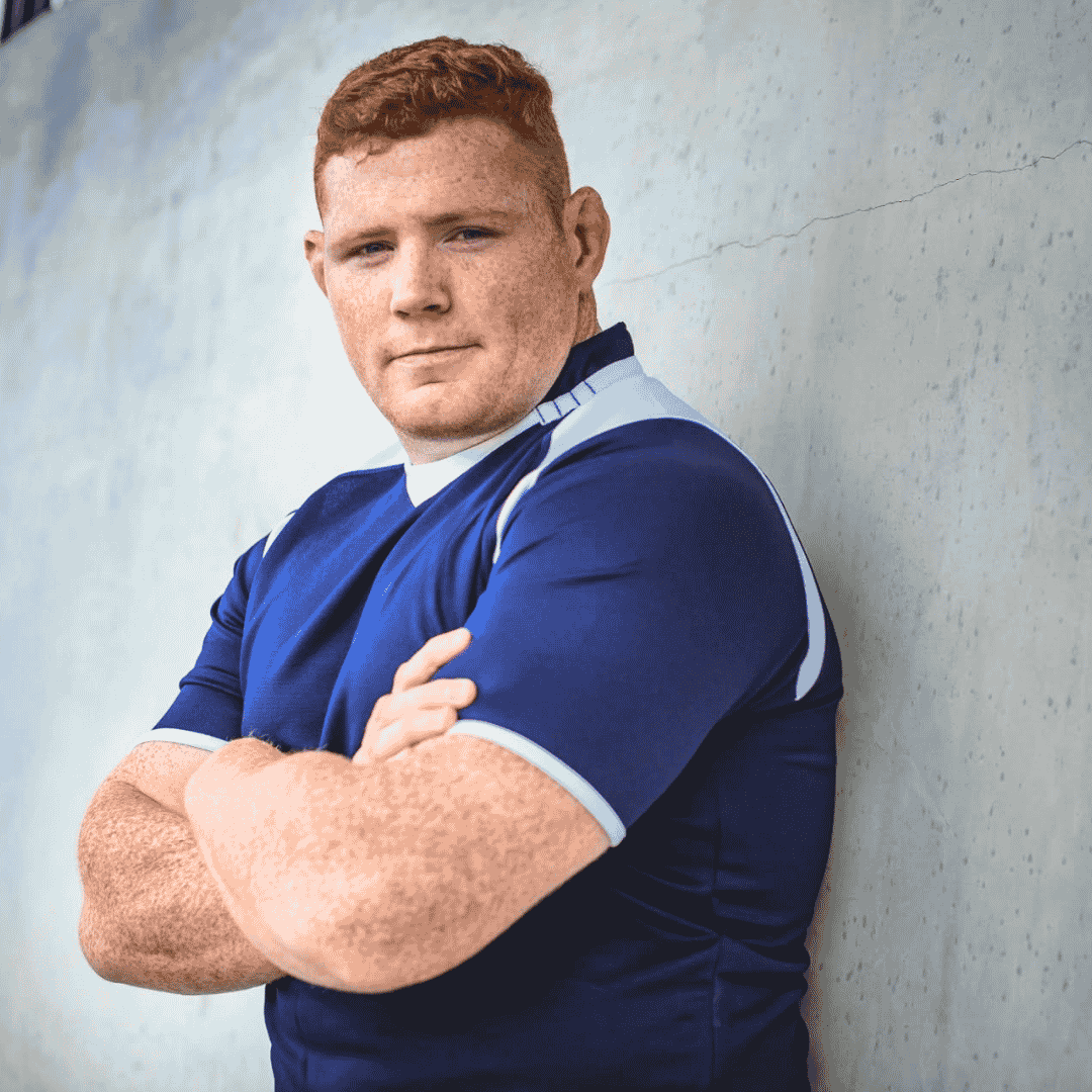 A young man with short red hair wearing a blue sports jersey stands with his arms crossed, leaning against a light-colored wall, looking confidently at the camera.