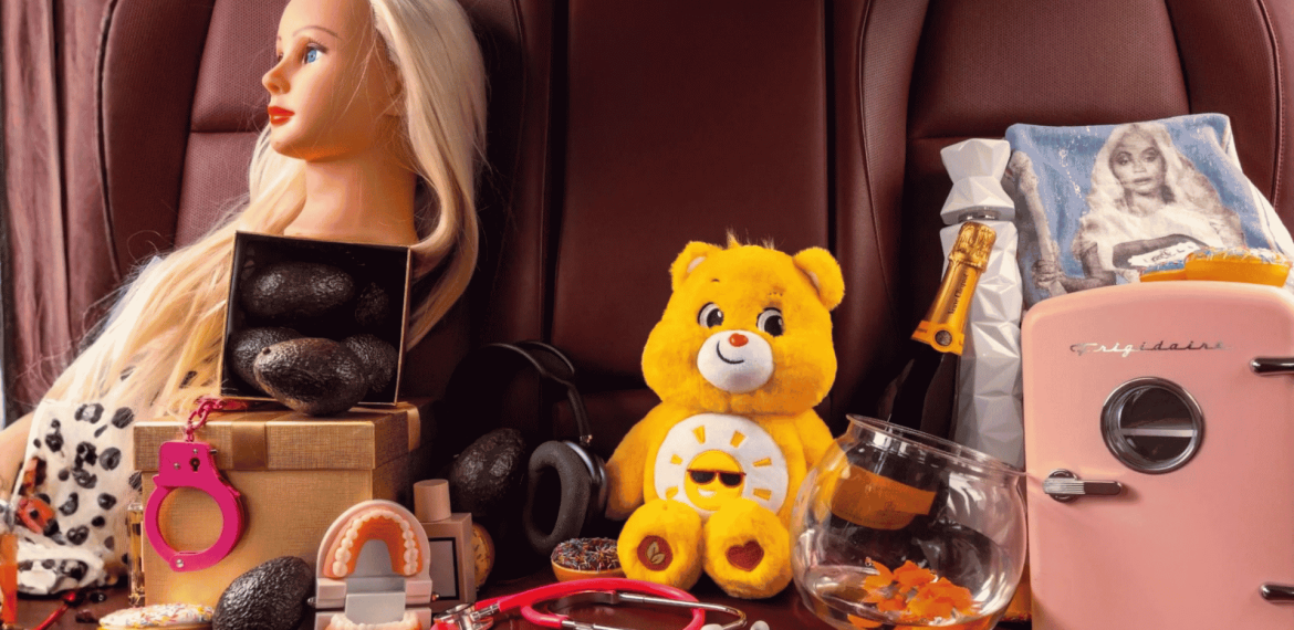 A collection of eclectic items on a leather seat, including a mannequin head, avocados, pink handcuffs, a teddy bear, a stethoscope, headphones, dental model, gift boxes, a glass bowl—a scene worthy of an Ali Campbell album cover.