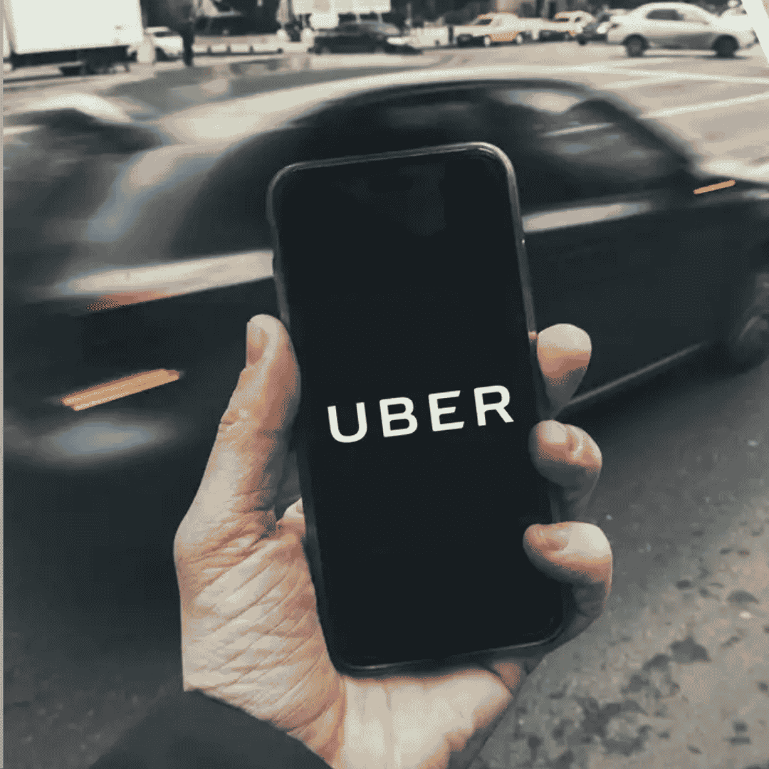 A hand holds a smartphone displaying the Uber logo, while a car blurs by on a city street—capturing the fast-paced vibe Ali Campbell might sing about.