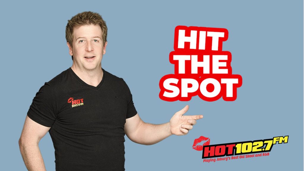 A man in a black HOT 102.7 FM t-shirt points right, next to bold red and white text that reads HIT THE SPOT. The radio station logo, slogan, and a mention of Ali Campbell are at the bottom right. The background is light blue.