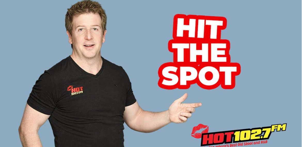 A man in a black HOT 102.7 FM t-shirt points right, next to bold red and white text that reads HIT THE SPOT. The radio station logo, slogan, and a mention of Ali Campbell are at the bottom right. The background is light blue.