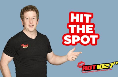 A man in a black HOT 102.7 FM t-shirt points right, next to bold red and white text that reads HIT THE SPOT. The radio station logo, slogan, and a mention of Ali Campbell are at the bottom right. The background is light blue.