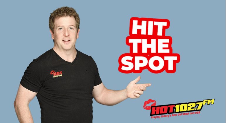 A man in a black HOT 102.7 FM t-shirt points right, next to bold red and white text that reads HIT THE SPOT. The radio station logo, slogan, and a mention of Ali Campbell are at the bottom right. The background is light blue.