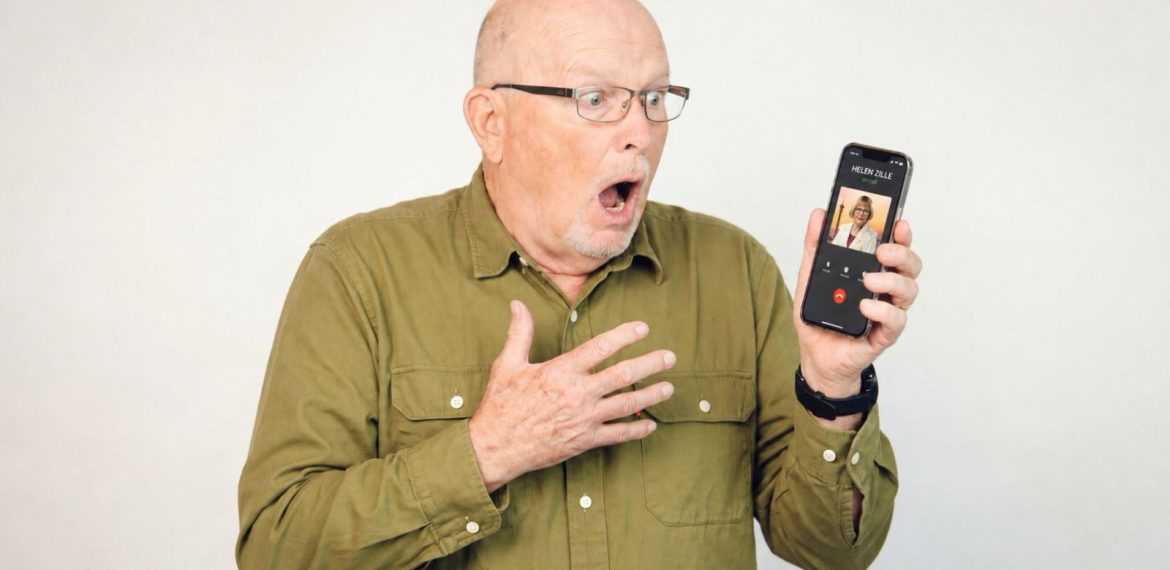 An older man wearing glasses and a green shirt looks surprised and holds a smartphone showing an incoming video call. His mouth is open and one hand is on his chest, expressing shock.