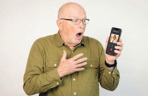 An older man wearing glasses and a green shirt looks surprised and holds a smartphone showing an incoming video call. His mouth is open and one hand is on his chest, expressing shock.