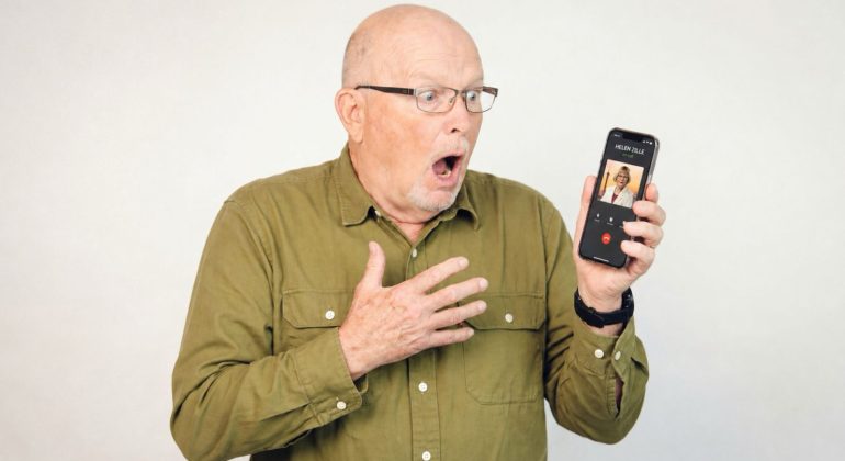 An older man wearing glasses and a green shirt looks surprised and holds a smartphone showing an incoming video call. His mouth is open and one hand is on his chest, expressing shock.