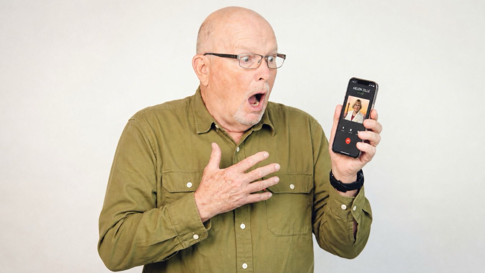 An older man wearing glasses and a green shirt looks surprised and holds a smartphone showing an incoming video call. His mouth is open and one hand is on his chest, expressing shock.