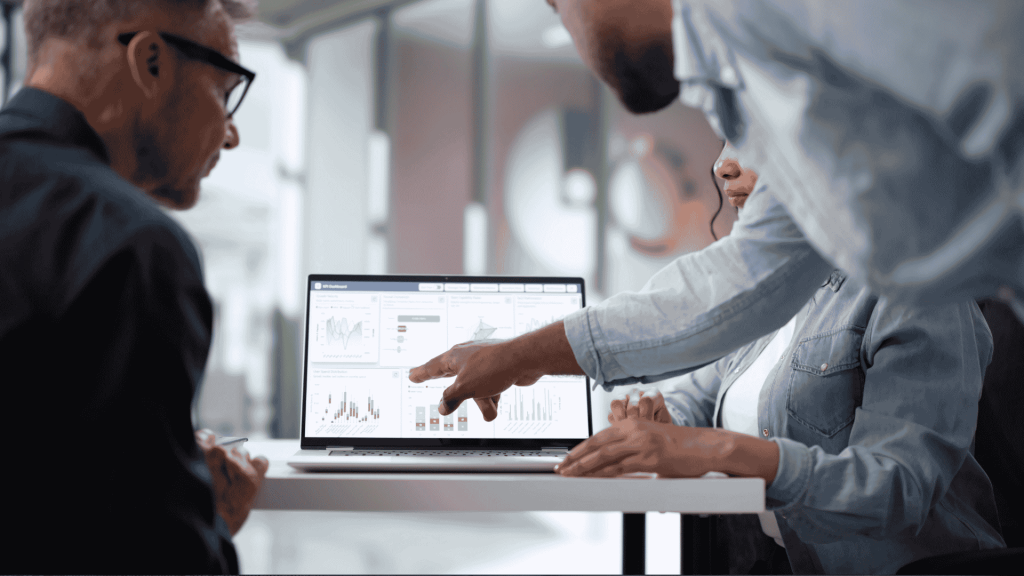 Three people sit at a table looking at a laptop displaying charts and graphs. One person points at the screen, discussing the data, while the others observe attentively in a modern office setting.