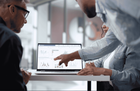 Three people sit at a table looking at a laptop displaying charts and graphs. One person points at the screen, discussing the data, while the others observe attentively in a modern office setting.