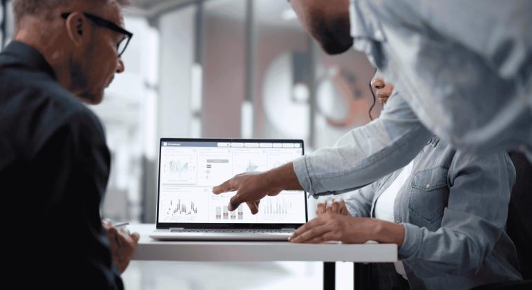 Three people sit at a table looking at a laptop displaying charts and graphs. One person points at the screen, discussing the data, while the others observe attentively in a modern office setting.