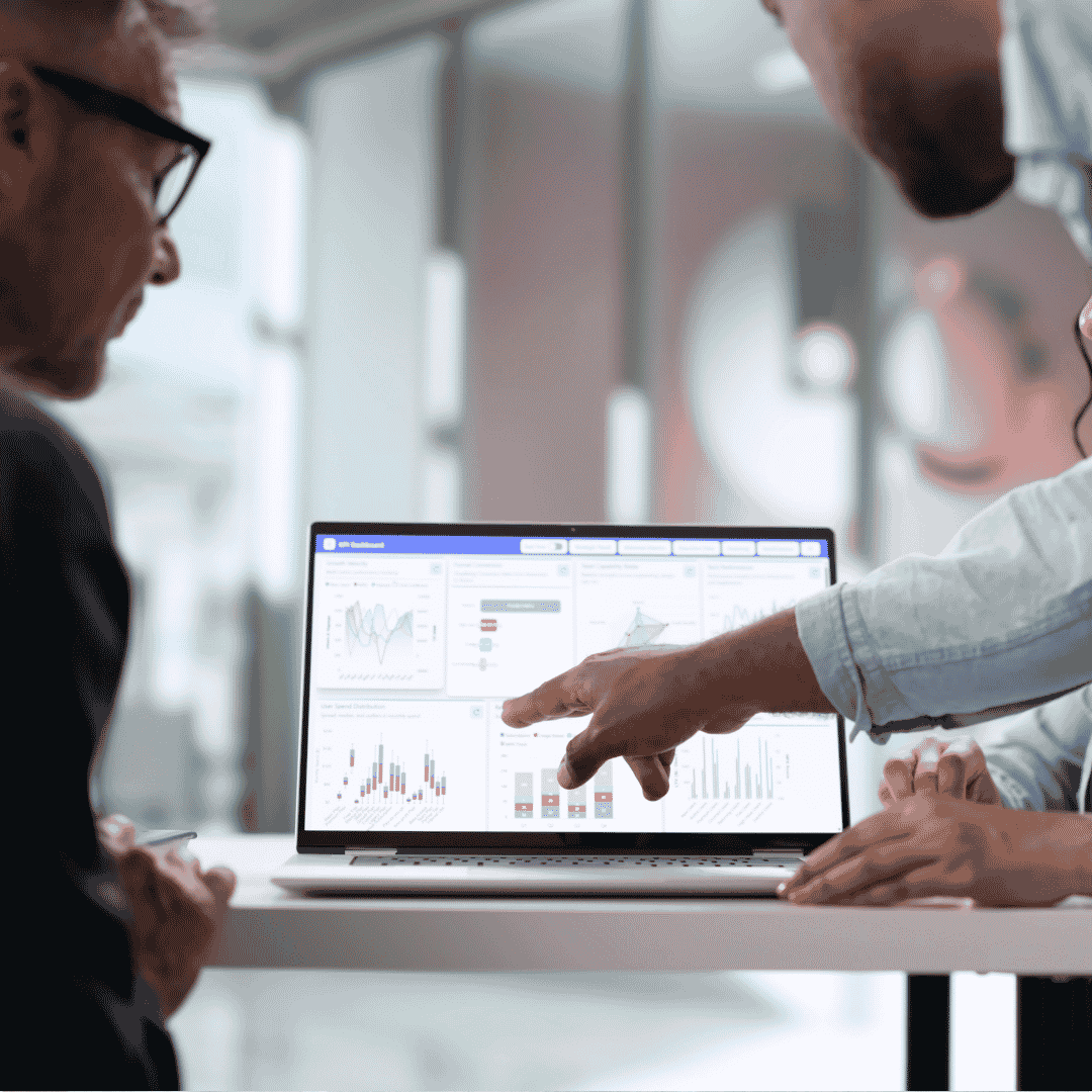 Three people sit at a table, looking at a laptop displaying data dashboards with charts and graphs. Two individuals are pointing at the screen, discussing the analytics shown.