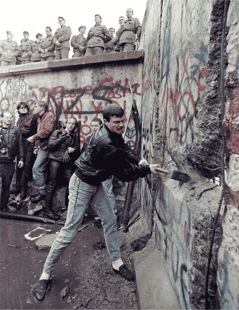 A man swings a sledgehammer at the Berlin Wall as a crowd watches, some standing atop the wall. The wall is covered in graffiti, symbolizing the fall of the Berlin Wall.
