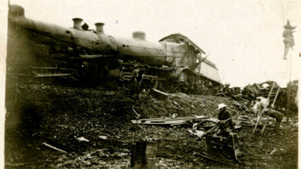 A damaged steam locomotive lies on its side amid scattered debris, possibly after experiencing one of the coldest temperature drops. Several people stand nearby, examining the black-and-white wreckage and working at the scene.