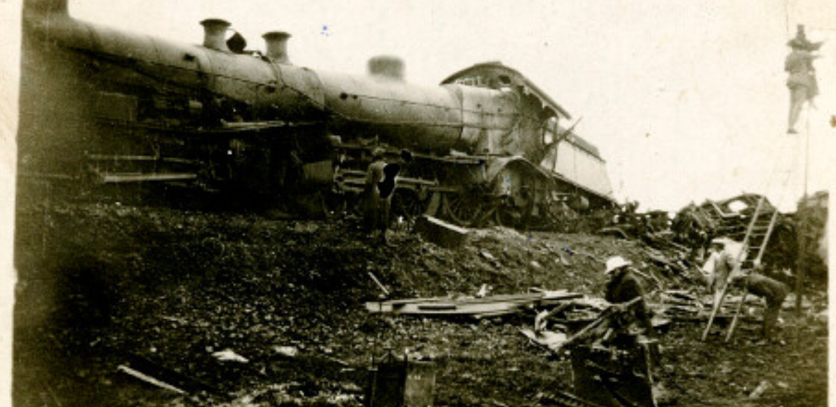 A damaged steam locomotive lies on its side amid scattered debris, possibly after experiencing one of the coldest temperature drops. Several people stand nearby, examining the black-and-white wreckage and working at the scene.