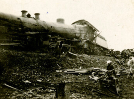 A damaged steam locomotive lies on its side amid scattered debris, possibly after experiencing one of the coldest temperature drops. Several people stand nearby, examining the black-and-white wreckage and working at the scene.