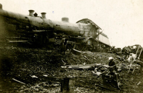 A damaged steam locomotive lies on its side amid scattered debris, possibly after experiencing one of the coldest temperature drops. Several people stand nearby, examining the black-and-white wreckage and working at the scene.