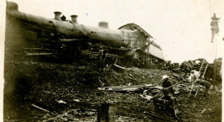 A damaged steam locomotive lies on its side amid scattered debris, possibly after experiencing one of the coldest temperature drops. Several people stand nearby, examining the black-and-white wreckage and working at the scene.