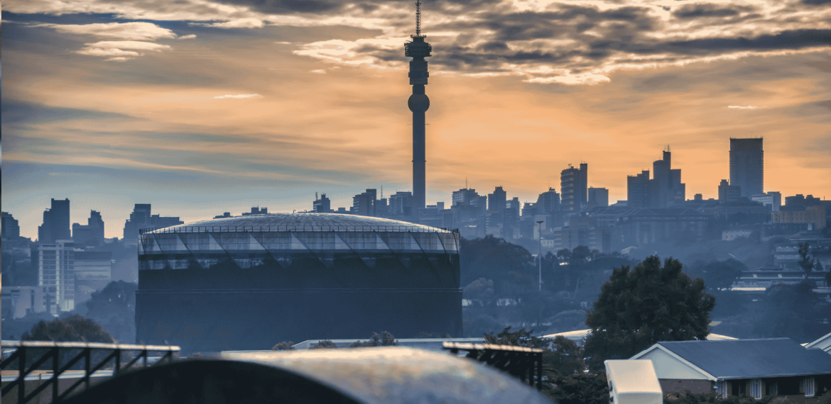 Johannesburg city skyline at sunset with the Hillbrow Tower prominent in the center, silhouetted buildings, and dramatic clouds in the sky.