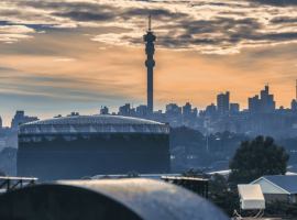 Johannesburg city skyline at sunset with the Hillbrow Tower prominent in the center, silhouetted buildings, and dramatic clouds in the sky.